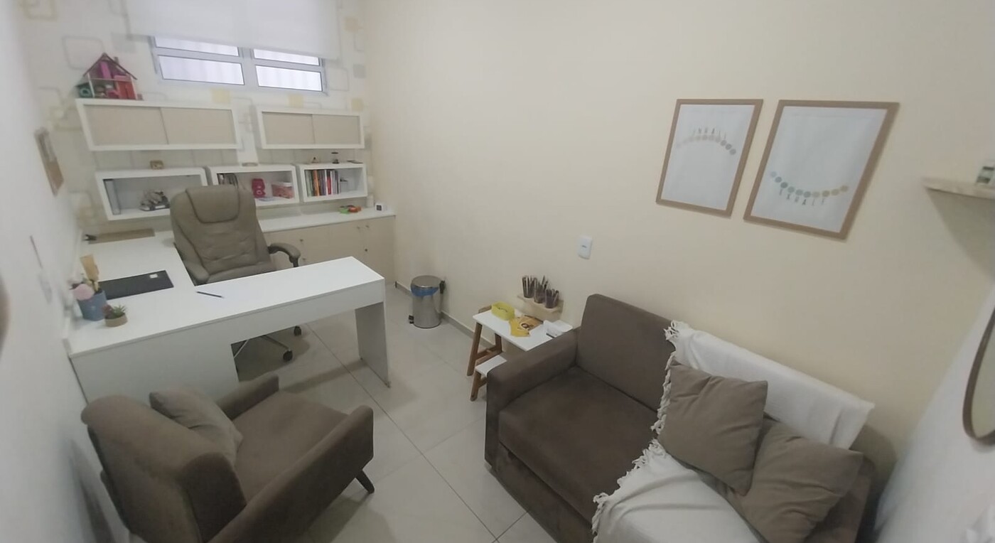 Sala de atendimento com mesa, poltrona e sofá, com quadros na parede e cantinho lúdico para crianças